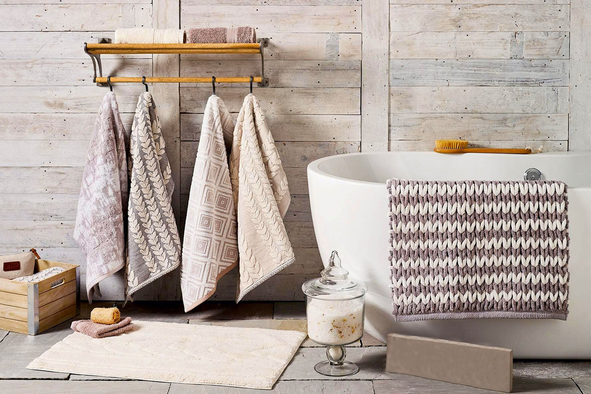 A cozy bathroom scene with a freestanding bathtub adorned with a textured towel. Various towels hang on a rustic rack against a wooden wall. A glass jar with bath salts and a wooden brush are nearby. A wooden box holds toiletries, and a bath mat lies on the floor.