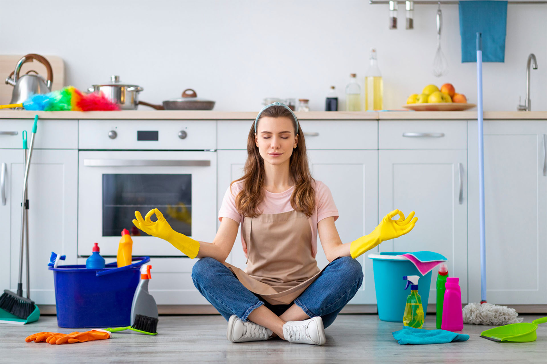 A woman sits cross-legged on a kitchen floor in a meditative pose, wearing yellow cleaning gloves. Cleaning supplies, including a mop, bucket, and bottles, are around her. The kitchen is white with a countertop holding various items.