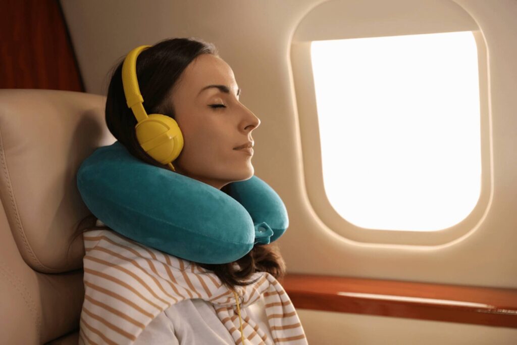 A person wearing yellow headphones and a blue neck pillow enjoys Jet-Set Bliss, sleeping while seated on an airplane. Natural light filters through the window, enhancing the serene in-flight experience.