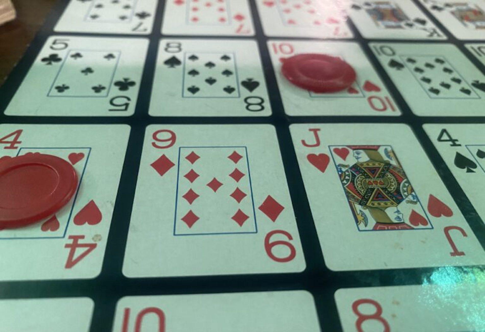 A board game featuring a grid of playing card images, including the 4 of clubs, 8 of spades, 9 of diamonds, and Jack of hearts, is shown. Red plastic chips are placed on some of the cards.