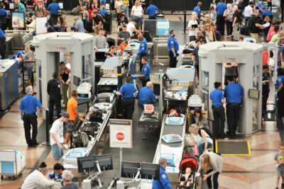 A busy airport security checkpoint buzzes with travelers efficiently moving through screening. Security personnel oversee the process as people prepare their belongings for X-ray machines. Some enjoy stress-free travel by saving time in line, while others collect items from conveyor belts.