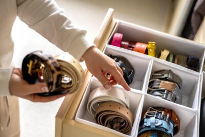 A chic busy woman is organizing a drawer with rolled-up belts of various colors and styles. The belts are neatly arranged in compartments, and she holds a couple of them in her hands, ready to add the final touches to her stylish collection.