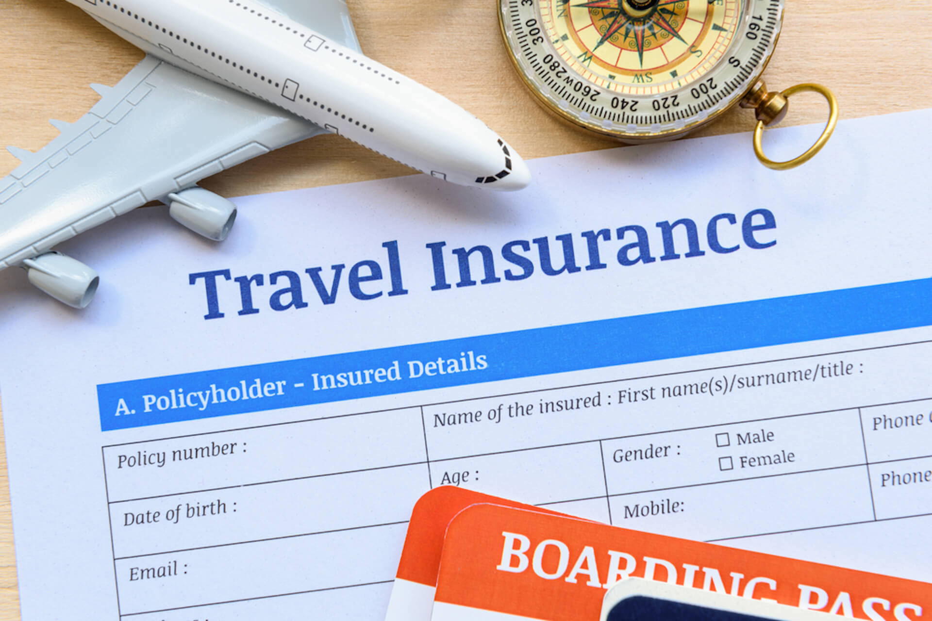 A paper with Travel Insurance printed on it lies under a toy airplane, a compass, and a boarding pass—essentials for avoiding flight fiascos. The document includes fields for policyholder information, such as name, age, and gender.