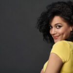 A person with curly hair and a playful smile is looking over their shoulder, exuding an air of romantic chemistry in a yellow shirt. The background is a solid dark grey.