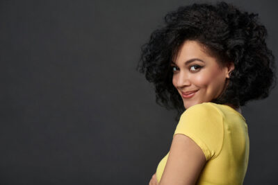 A person with curly hair and a playful smile is looking over their shoulder, exuding an air of romantic chemistry in a yellow shirt. The background is a solid dark grey.