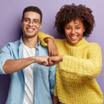 A smiling man and woman stand in front of a purple background, wearing casual clothes. Fist-bumping with enthusiasm, they celebrate the positive connection that comes from working together in harmony.