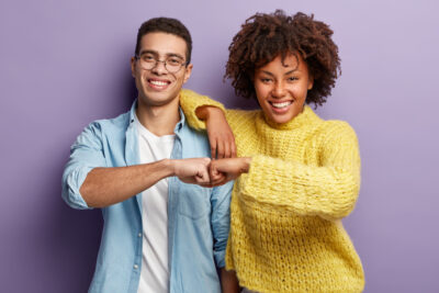 A smiling man and woman stand in front of a purple background, wearing casual clothes. Fist-bumping with enthusiasm, they celebrate the positive connection that comes from working together in harmony.