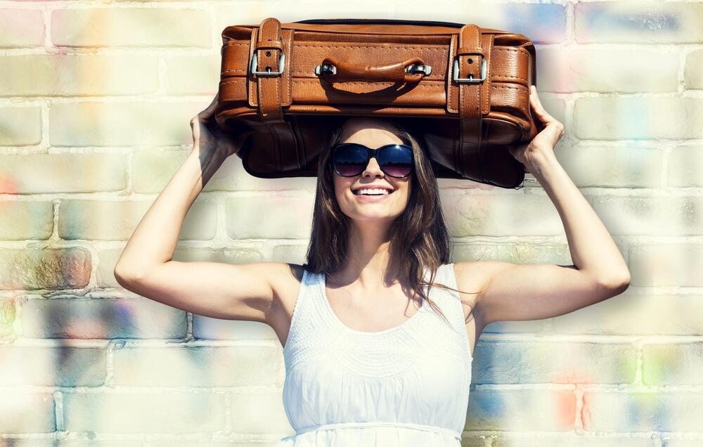 A smiling woman in sunglasses and a white dress holds a brown suitcase above her head, ready for a Girls' Trip Adventure. She stands in front of a colorful, graffiti-covered brick wall, capturing the spirit of fun and excitement.