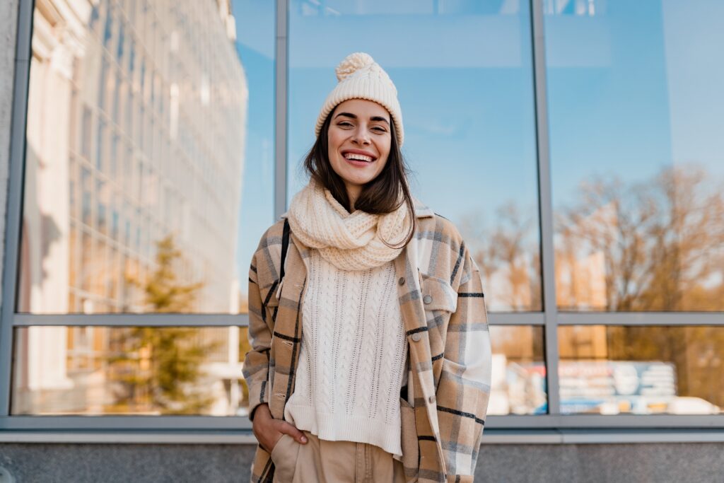 A smiling woman wearing a beige beanie, scarf, and plaid jacket stands in front of a building with large windows. The reflection shows a clear blue sky and trees, adding a touch of stylish winter charm to the urban setting. Embrace the chill with frosty fashion finds this season.