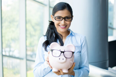 A smiling woman with dark hair and glasses stands indoors, holding a piggy bank also wearing glasses. Dressed in a light blue shirt, she embodies the savvy art of staying rich. The background features large windows with a blurred view outside.