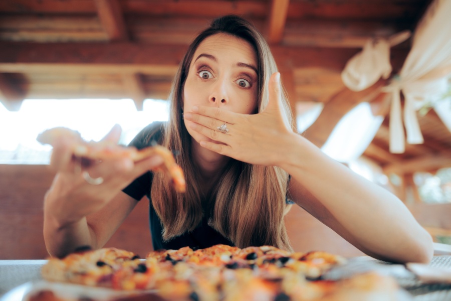 A surprised woman with long brown hair holds a slice of pizza in one hand and covers her mouth with the other, pondering food swaps. She's sitting at a table with more pizza, in a wooden outdoor setting.