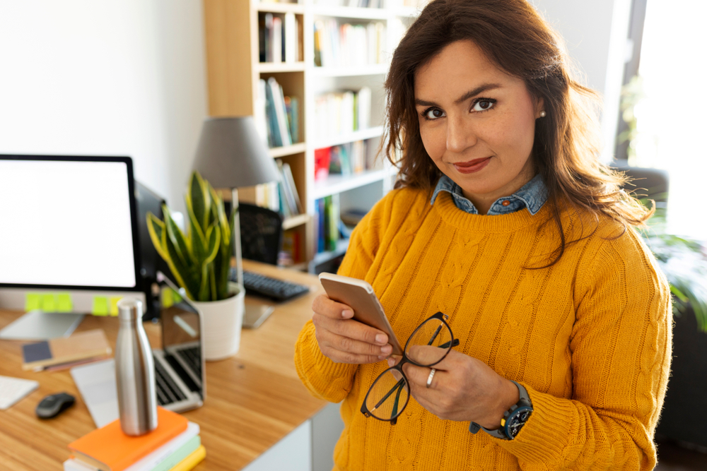 A woman in a yellow sweater, embracing remote work, holds a smartphone and eyeglasses while standing in her home office. Behind her is a desk with a computer, books, and potted plant. She smiles slightly at the camera, showcasing her well-organized remote workspace.