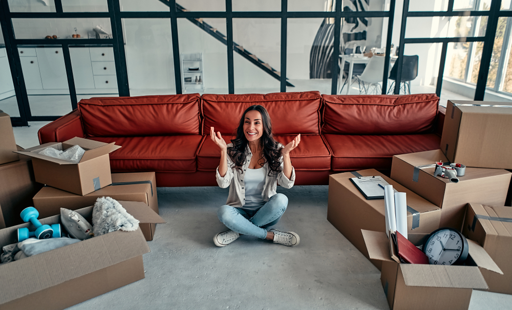 A woman sits on the floor with her arms up, surrounded by open moving boxes in a bright, modern condo. A red sofa and large glass windows are in the background, reflecting her secure investment in a space filled with unpacked items.