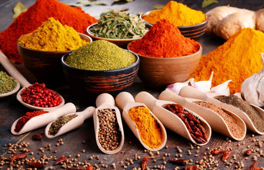 An assortment of colorful spices displayed in bowls and wooden scoops. Includes turmeric, chili powder, green herbs, and peppercorns, arranged on a wooden surface for a vibrant and aromatic presentation that inspires flavorful creations.