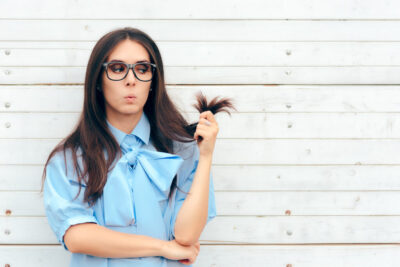 A woman with long dark hair and glasses stands against a light wooden wall. Clad in a blue blouse with a large bow, she playfully holds a section of her hair while pouting thoughtfully, as though considering how to transform her tresses and banish split ends for good.