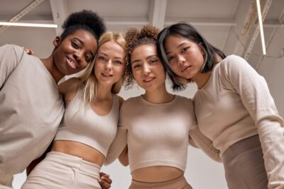 Four women stand close together, smiling, with their arms around each other. They are dressed in light-colored tops and showcase different hair types, enhancing the vibrant atmosphere of the bright indoor space.