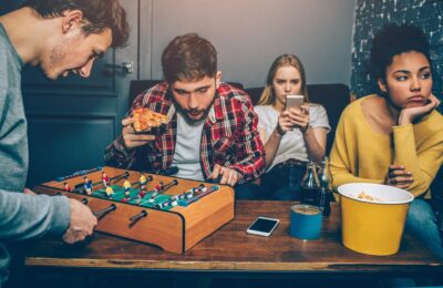 In a casual room, four people enjoy a relaxed evening. Two are playing foosball while one munches pizza and another checks their phone. A popcorn bucket and drinks on the table enhance the laid-back vibe, making it the perfect setting to deal with friends or share relationship advice.