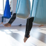 People practicing aerial yoga in a bright studio, suspended in blue hammocks with their legs and feet visible. Sunlight streams through large windows, illuminating the floor, creating a serene zen ambiance as they appear to be flying high above the ground.