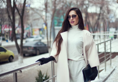 A woman from the Busy Beauties collection poses in stylish winter attire, including sunglasses, a white coat, and black gloves. She stands outdoors against a snowy urban backdrop with parked cars and leafless trees, embodying the elegance of winter chic.