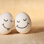 Two eggs with drawn faces rest on a wooden surface, strengthening their bond. The egg on the left sports closed eyes, a smile, and its tongue sticking out; the egg on the right beams with a wide smile. These playful friends capture the essence of togetherness and camaraderie.