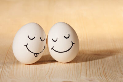 Two eggs with drawn faces rest on a wooden surface, strengthening their bond. The egg on the left sports closed eyes, a smile, and its tongue sticking out; the egg on the right beams with a wide smile. These playful friends capture the essence of togetherness and camaraderie.