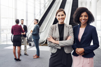 Two professional women stand confidently in the foreground with their arms crossed, smiling, exuding the spirit of an Entrepreneur Conference. In the background, connections are being made as three people engage in conversation near an escalator in a modern building with large glass windows.