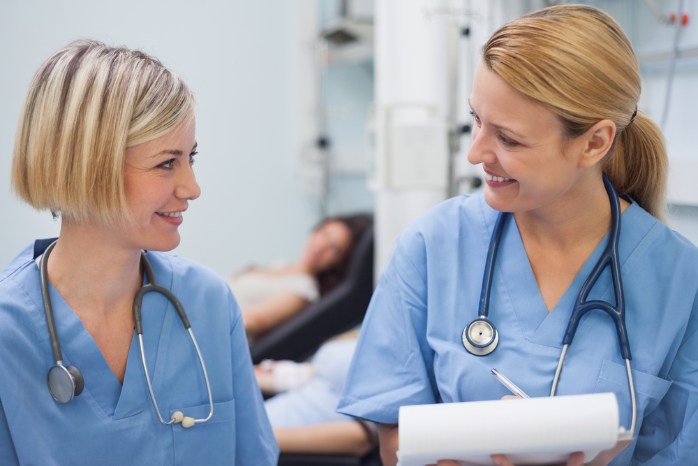 Two smiling healthcare professionals in blue scrubs and stethoscopes exchange tips while holding a clipboard. In the background, a patient rests on a bed in a light-colored medical room, benefiting from their collaborative feedback.