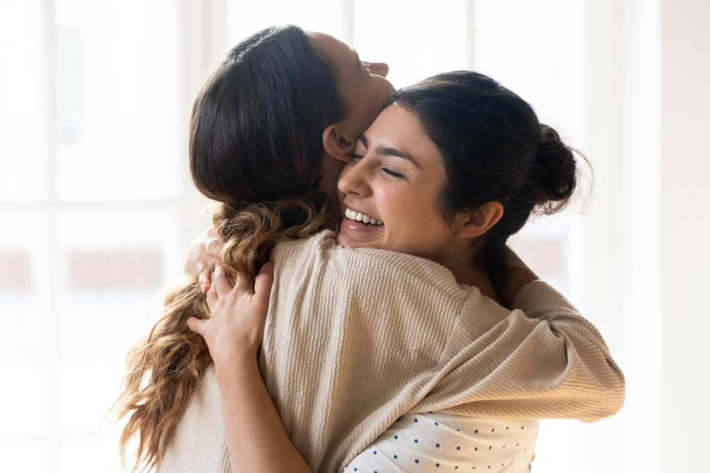 Two people hugging and smiling warmly in a sunlit room. The person facing the camera has dark hair pulled into a bun, while the other has long, wavy hair. Their embrace reflects a trusting relationship, conveying happiness and comfort as they rebuild their bond with affection.