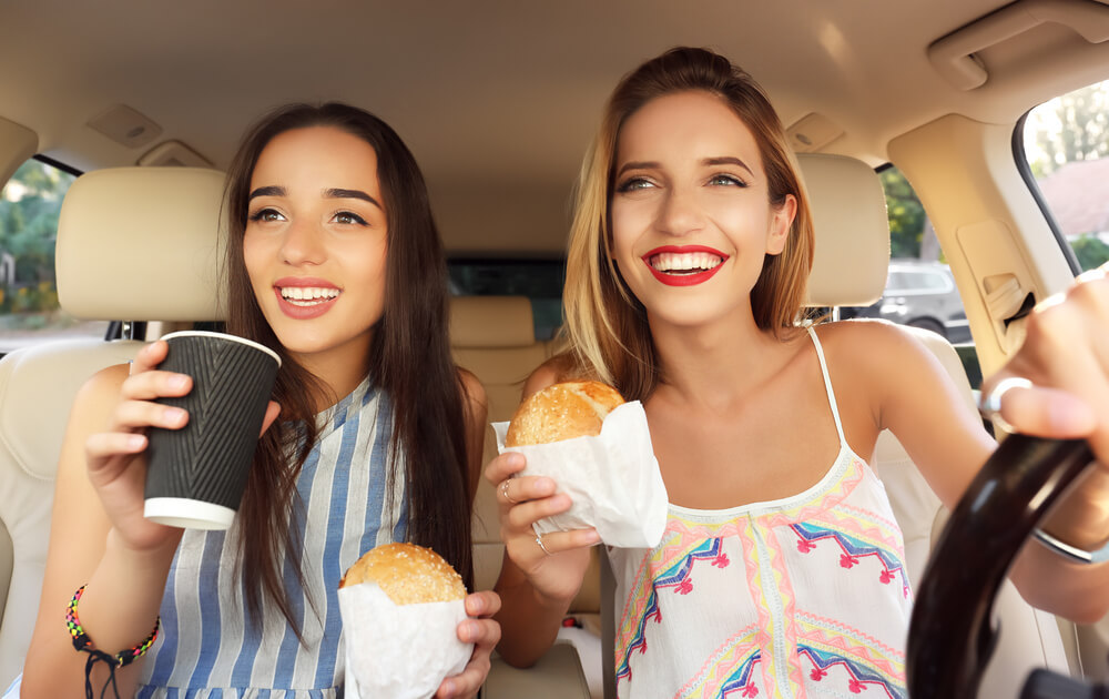 Two women sit in a car, smiling. The driver holds a steering wheel and a sandwich, while the passenger holds a sandwich and a paper cup. They appear to be enjoying their snacks during a road trip, with sunlight streaming through the windows.