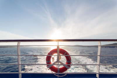 View from the back of a boat with a red life preserver attached to the railing. The sun sets over the ocean, casting reflections on the water. Distant land is visible on the horizon, epitomizing affordable cruising at its finest.