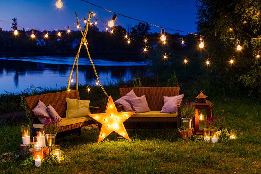 A cozy outdoor patio by a lake at dusk, featuring wicker sofas with purple cushions. String lights and mood lighting create an enchanting ambiance, enhanced by a glowing star-shaped lantern on the ground, surrounded by candles and potted plants, creating a warm and inviting atmosphere.