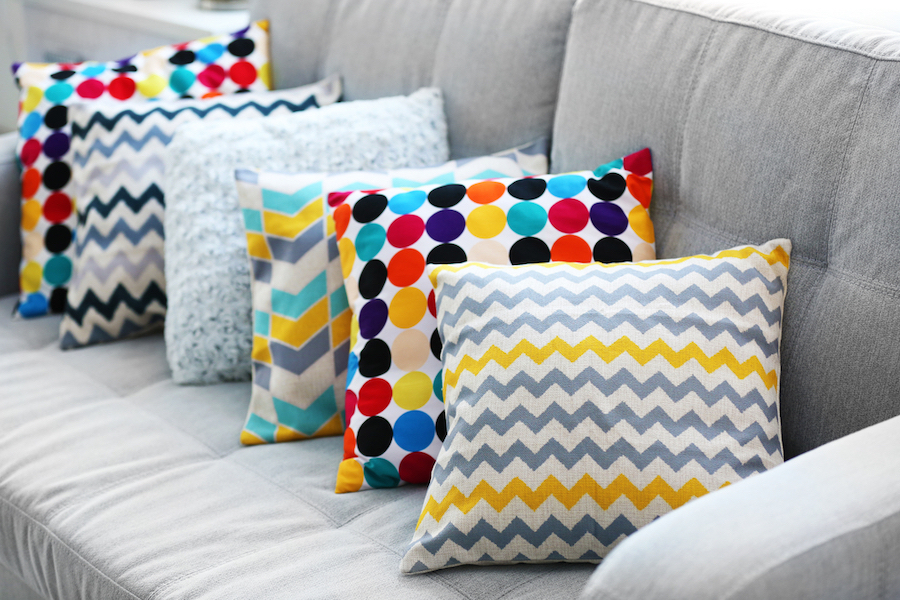 A gray sofa adorned with five throw pillows enhances the living room style. The pillows, with vibrant polka dots, blue and yellow chevrons, and abstract designs on their pillow covers, create a bright and modern setting.