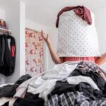 A person sits in a room overflowing with clothes, their head covered by a laundry basket. Their arms are raised in frustration from the closet organization project. The room is lined with racks of clothes and a floral-patterned curtain in the background, embodying chaos amid attempted order.