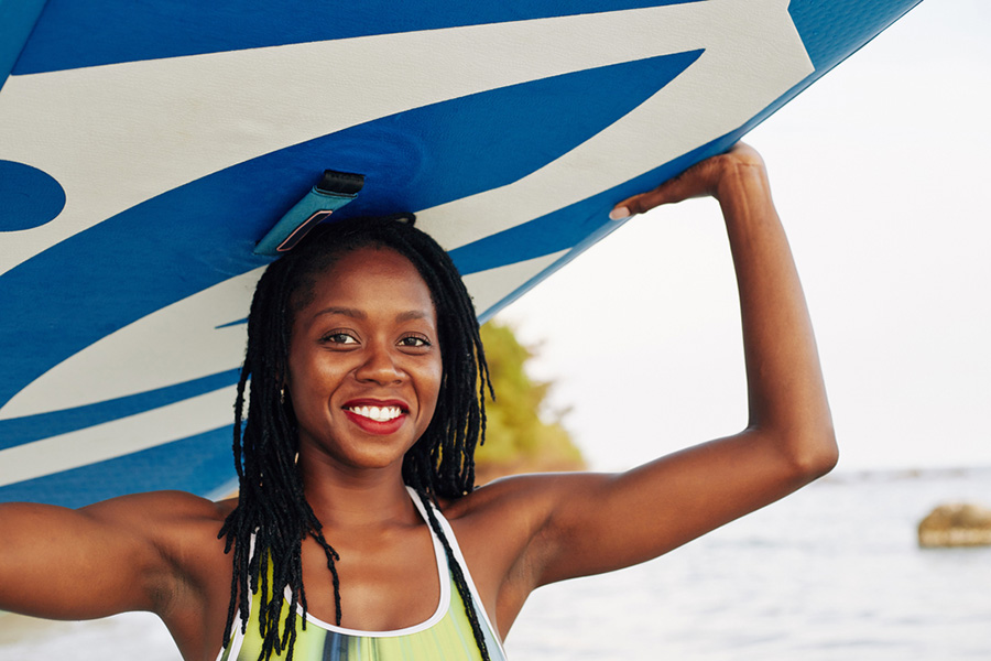 A smiling woman holds a blue paddleboard over her head near a body of water, embodying La Dolce Vita. She wears a colorful top and has long braided hair, embracing her Ageless Adventures. There's lush greenery visible in the background.