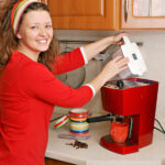 A woman with curly hair and a headband smiles while preparing coffee using a must-have red espresso machine in her kitchen's coffee station. Scattered coffee beans and colorful cups brighten the counter.