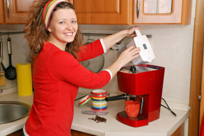 A woman with curly hair and a headband smiles while preparing coffee using a must-have red espresso machine in her kitchen's coffee station. Scattered coffee beans and colorful cups brighten the counter.