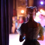 A ballerina stands backstage, silhouetted against the stage's bright lights, embodying theatre magic. She wears a floral headpiece and a lace costume, while other dancers blur by in a dazzling display of movement and light.