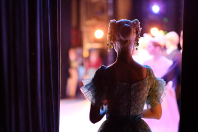 A ballerina stands backstage, silhouetted against the stage's bright lights, embodying theatre magic. She wears a floral headpiece and a lace costume, while other dancers blur by in a dazzling display of movement and light.