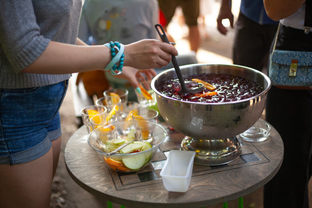 A person serving punch from a large bowl into cups at a lively girls brunch. The punch, enriched with fruit slices, sits beside additional glassware and a bowl of apples and oranges. Outdoors, surrounded by people, it's the perfect setting for sharing new punch recipes and enjoying delightful conversation.