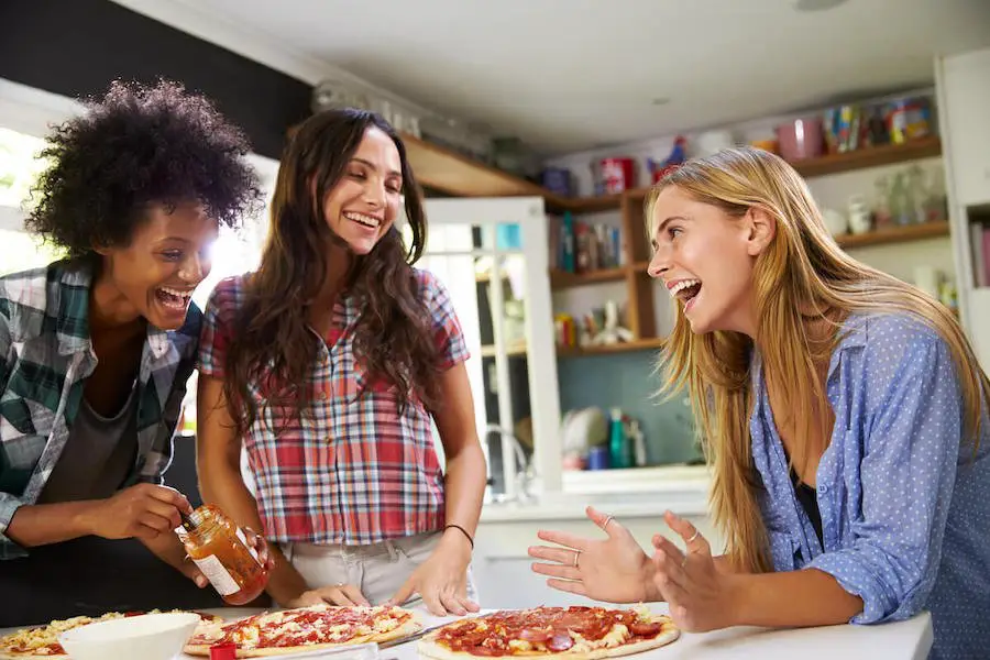 In a joyful and relaxed atmosphere, three women engage in a Pizza Bonding Party in the kitchen, laughing as one holds a jar of sauce while the others prepare toppings. Casually dressed, they enjoy each other's company with shelves stacked with unique finds like cups and plates in the background.