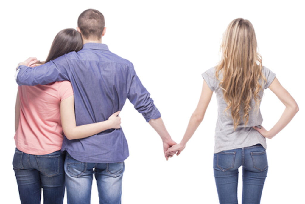 A man finds himself in a dating dilemma, standing between two women dressed casually. He wraps his arm around the woman on the left while holding hands with the woman on the right, all against a plain white background.