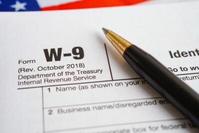 Close-up of a W-9 form from the U.S. Department of the Treasury, Internal Revenue Service, with a black pen resting on it. Part of the American flag is visible in the background.