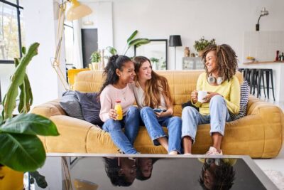 Three friends forge lifelong memories while sitting on a yellow couch in a modern living room, laughing and enjoying drinks. One holds a juice, another has coffee, and the third wears headphones around her neck. The room features lush plants and stylish decor, complementing their joyous camaraderie.