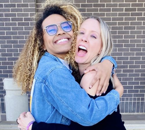 Two smiling individuals hugging outdoors, embodying the spirit of Busy Girl Nation. One with curly hair and glasses in a denim jacket, and the other with straight hair in a black top. They stand before a brick wall, radiating happiness and friendship.