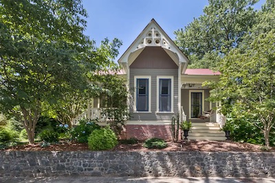 A charming, small house in Asheville, North Carolina, with a pointed roof and front porch, is surrounded by lush green trees and a stone wall. The exterior features light-colored siding and decorative trim under a clear blue sky—an ideal spot for enjoying local brews.