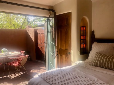 A cozy bedroom with sunlight streaming through open folding doors. The room features a neatly made bed with pillows and a textured blanket. Outside, there's a small patio with a round table, chairs, and a warm terracotta wall reminiscent of Santa Fe’s unique charm.