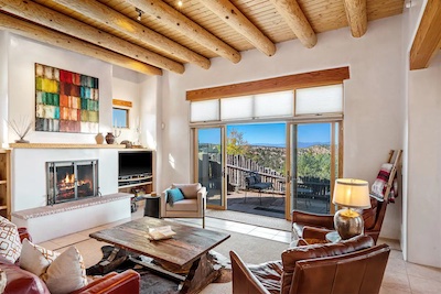 A cozy living room with a fireplace, wooden ceiling beams, and large windows overlooking a scenic outdoor patio. Infused with Santa Fe charm, the room features leather chairs, a wooden coffee table, and a colorful abstract painting above the fireplace.