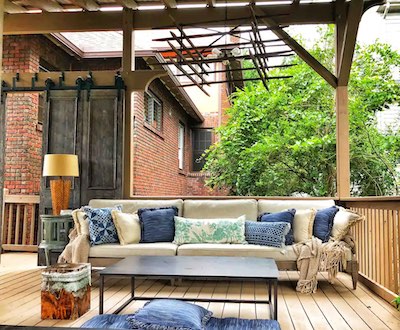 A cozy outdoor patio in Asheville features a large beige sectional sofa adorned with blue and patterned pillows. A wooden pergola provides shade, and there's a dark rectangular coffee table perfect for serving brews. Lush greenery peeks through the frame, complemented by a brick wall.