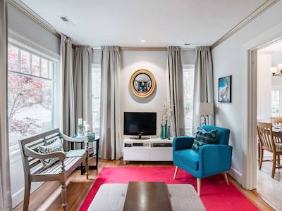 A cozy Portland living room features a turquoise armchair, a white TV stand with a small television, and a wooden bench with cushions on a red rug. Large windows with curtains flood the space with natural light, highlighting the round mirror on the wall and opening up endless possibilities.