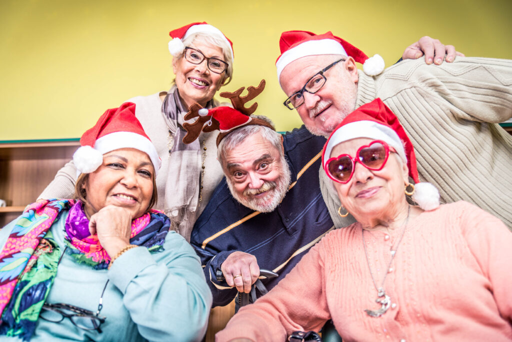 A group of five seniors, delightfully smiling and wearing festive Santa hats, gathers closely together. One dons reindeer antlers while another sports heart-shaped sunglasses, capturing the joy often found in retirement communities. They are set against a cheerful green backdrop.